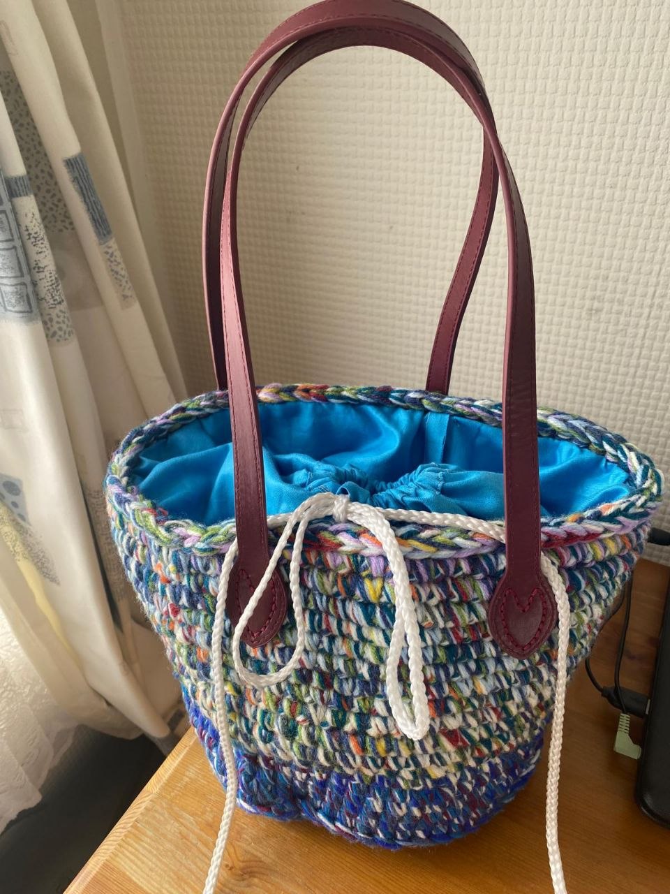 Colourful crochet basket bag with burgundy handles, blue lining, and white drawstring closure